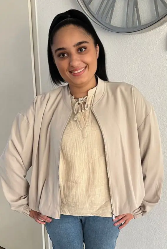 Young woman in a beige bomber jacket and light-colored top smiles.